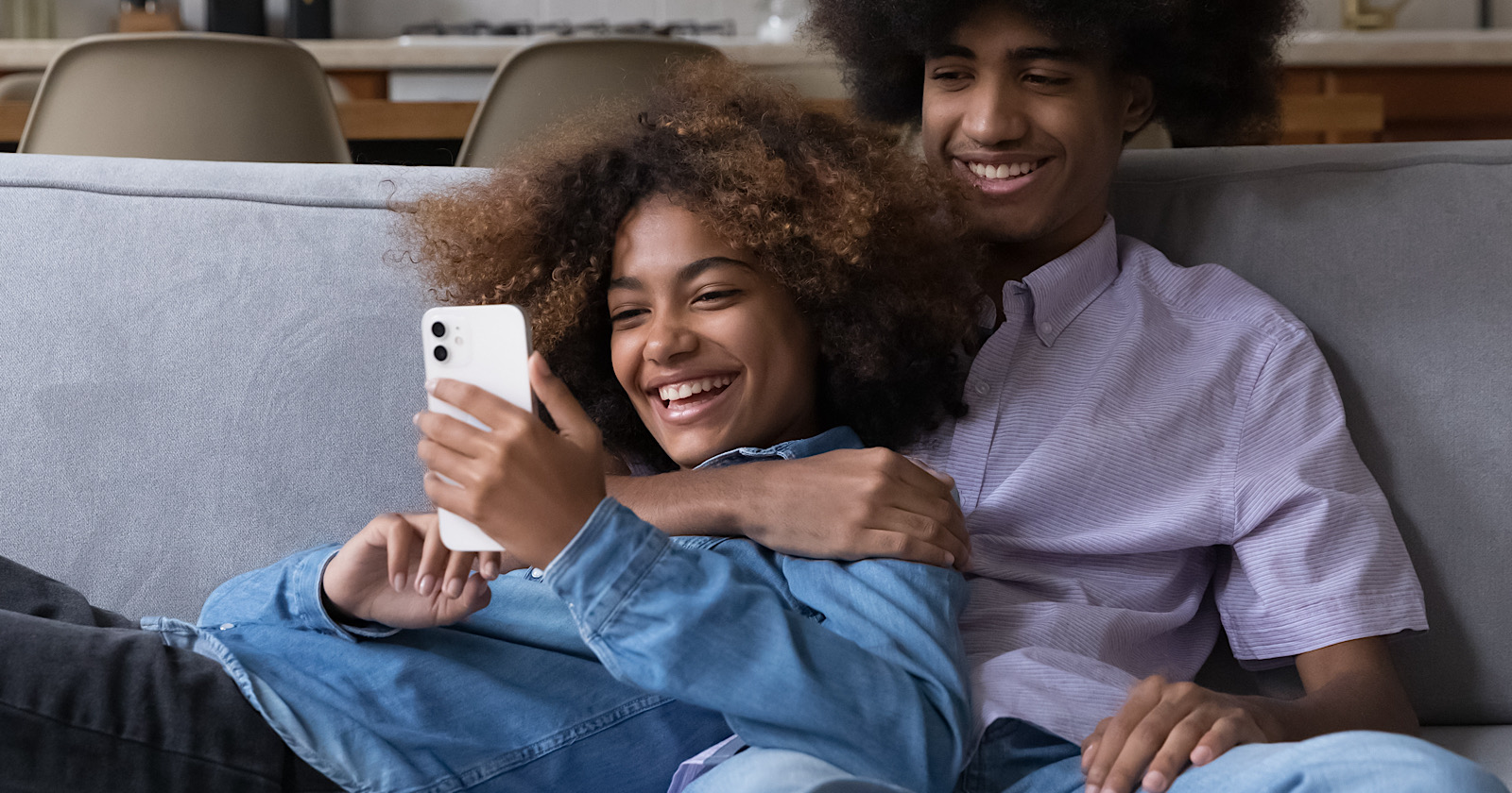 Smiling African teenager couple relaxing on sofa staring at cellphone screen