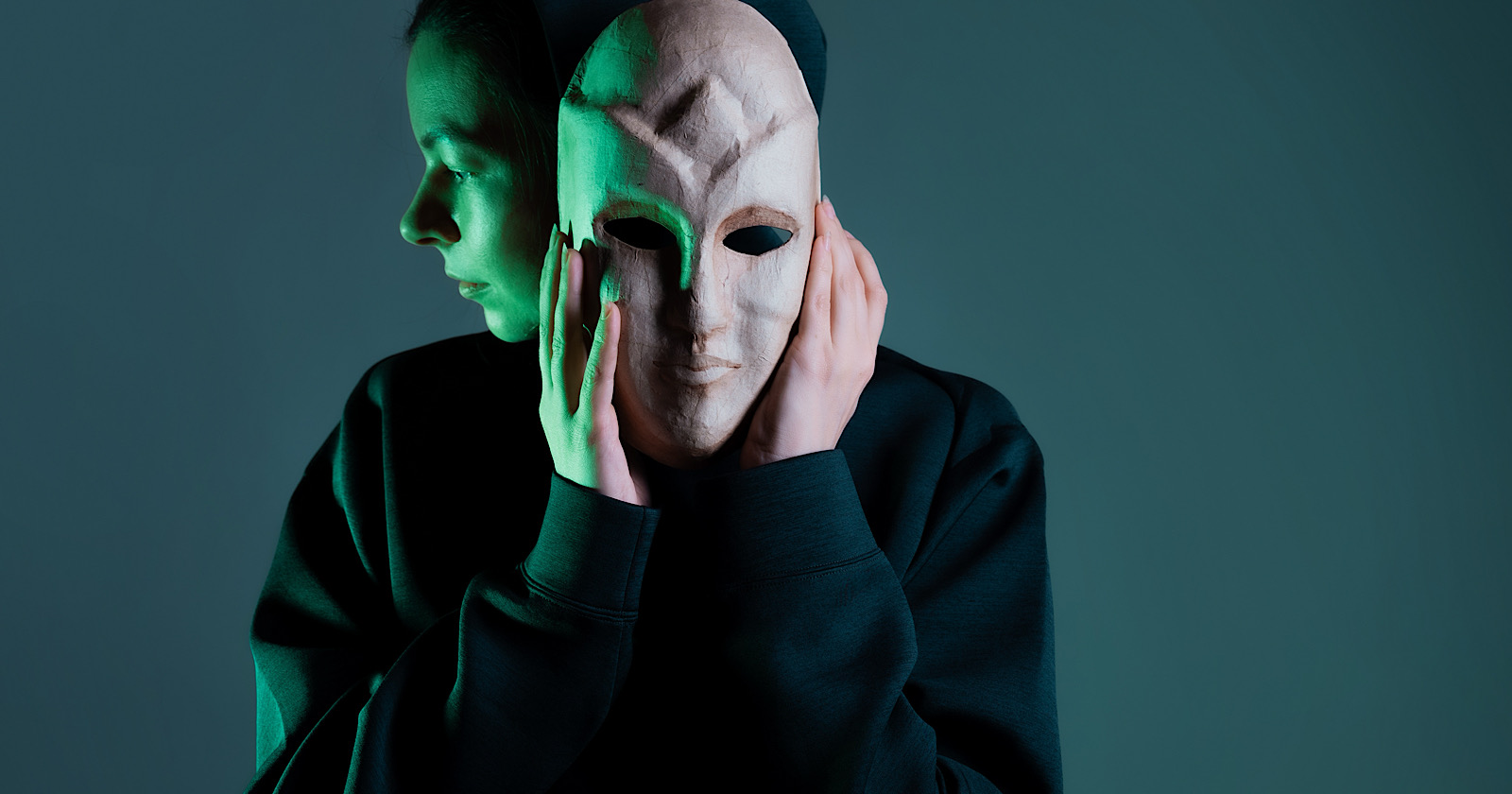 Hiding behind a mask, a young woman in a dark hoodie hides her face with a mask, privacy on the Internet, the concept of information security