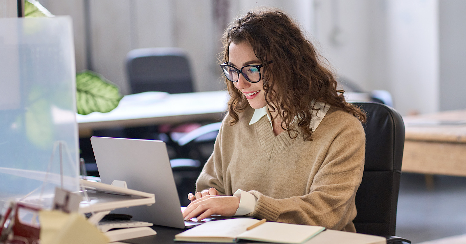 Young happy professional business woman employee sitting at desk working on laptop in modern corporate office interior. Smiling female worker using computer technology typing browsing web.