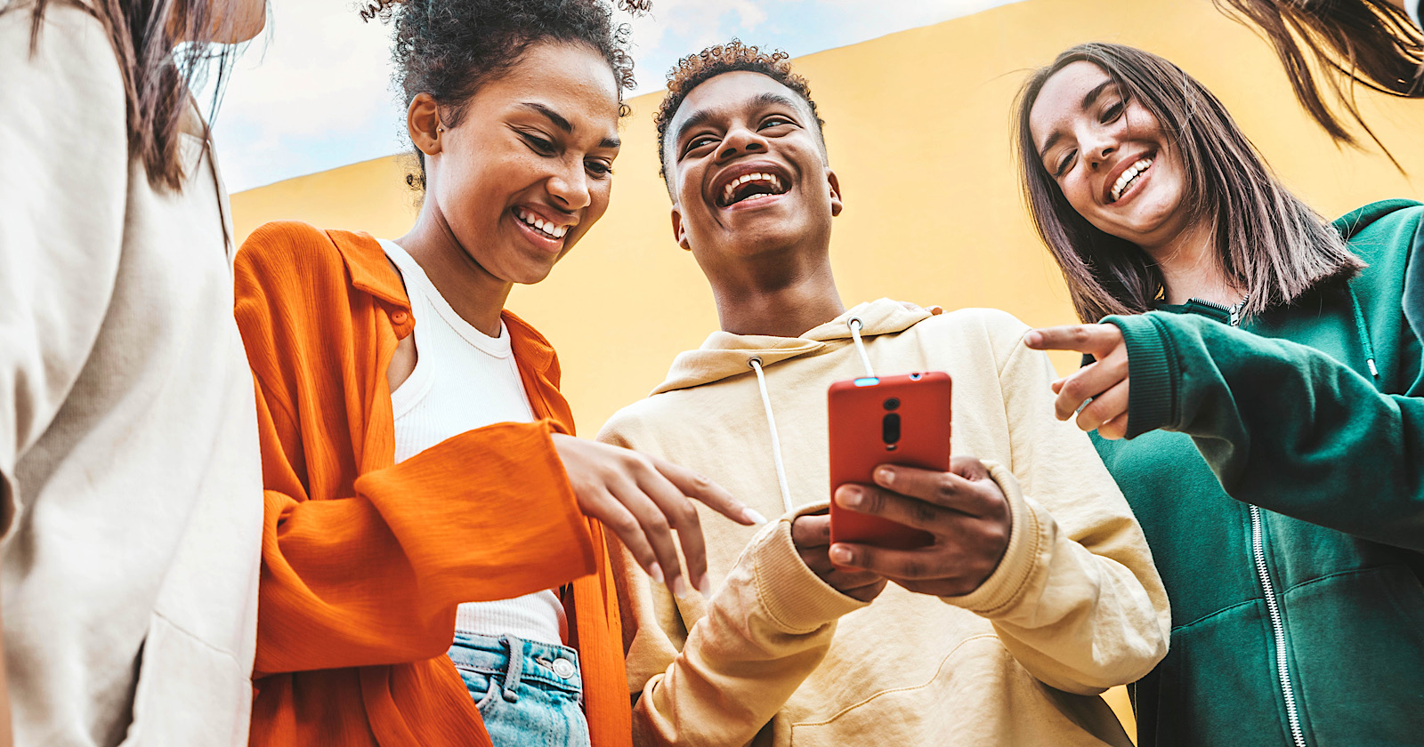 A divers  radical  of Gen Z friends laughing and looking astatine  a smartphone held by a young man, outdoors nether  a bright, colorful background.